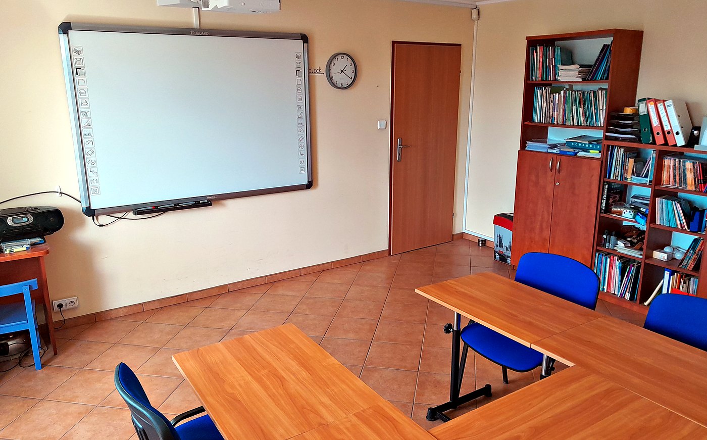Sala AcCentu: tablica interaktywna, zegar i regał z książkami przy stołach